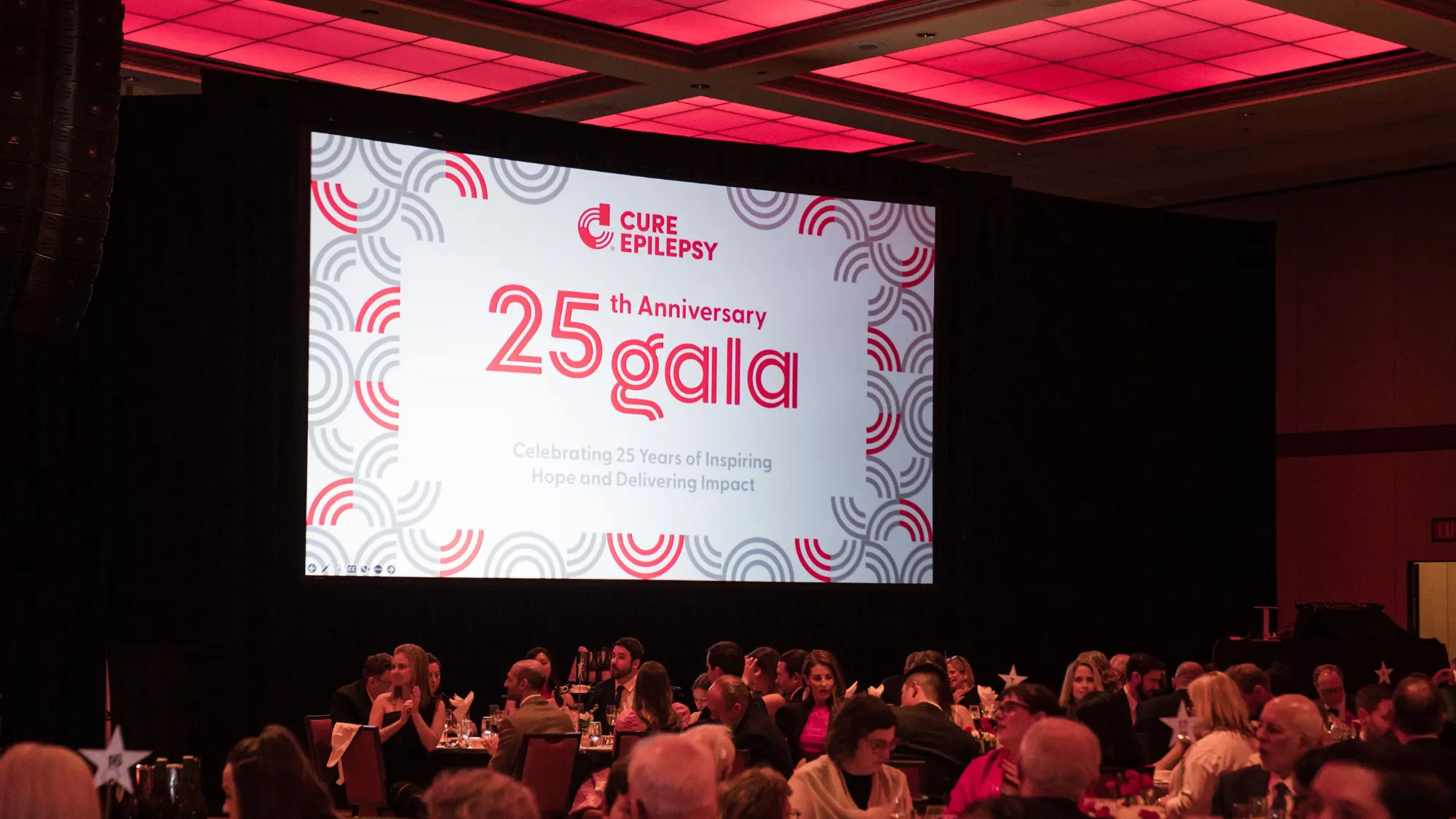 Photo of the CURE Epilepsy 25th Anniversary Gala event with the branding and logo on a large screen in front of an audience.