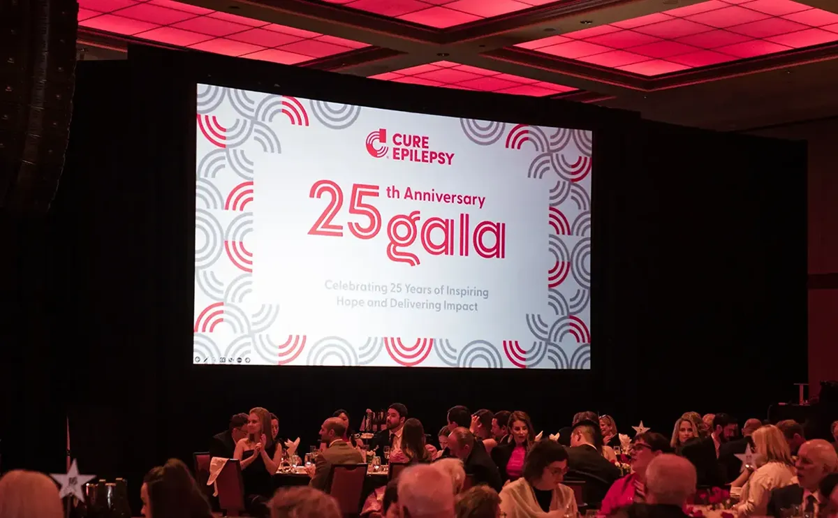 Photo of the CURE Epilepsy 25th Anniversary Gala event with the branding and logo on a large screen in front of an audience.