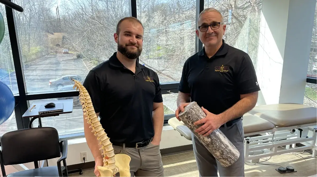 Two physical therapists posing with a foam roller and spine model.